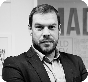 Portrait en noir et blanc d'un homme barbu portant un costume sombre et une chemise claire, regardant directement la caméra avec un léger sourire.