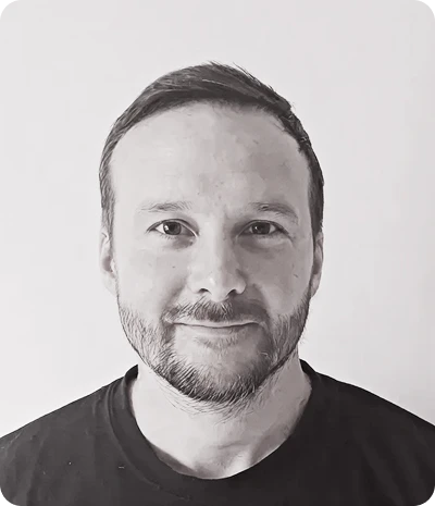 Portrait en noir et blanc d'un homme souriant avec une barbe légère et un t-shirt noir.