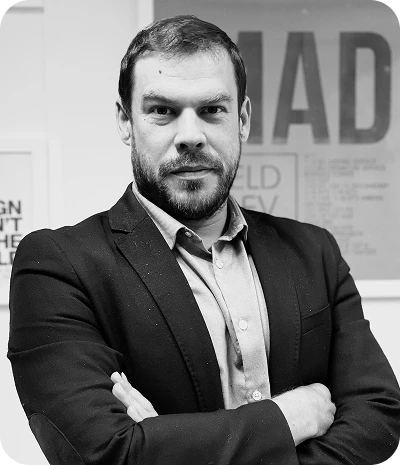 Homme aux cheveux courts et barbe portant une veste noire et chemise claire, bras croisés, regardant l'appareil photo.