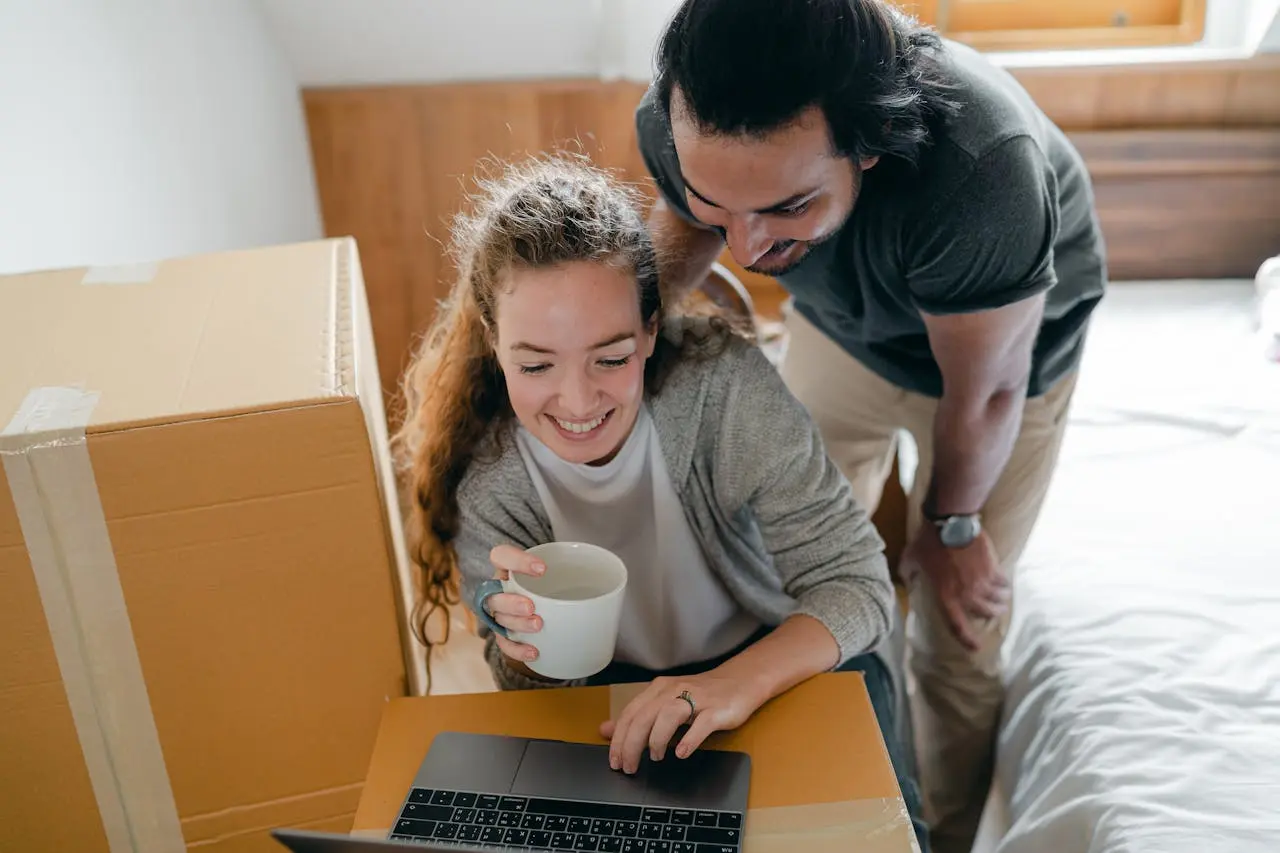 Un couple souriant regarde un ordinateur portable posé sur une boîte en carton, la femme tient une tasse blanche.