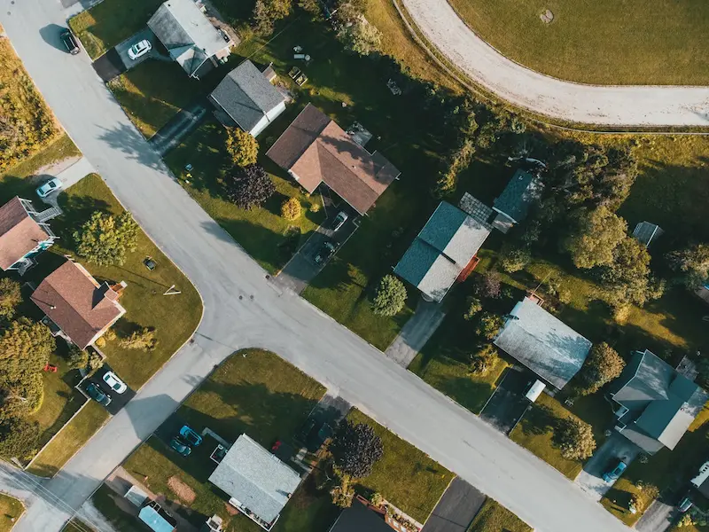 Vue aérienne d'un croisement de rues dans un quartier résidentiel avec des maisons, des pelouses vertes et quelques voitures stationnées.