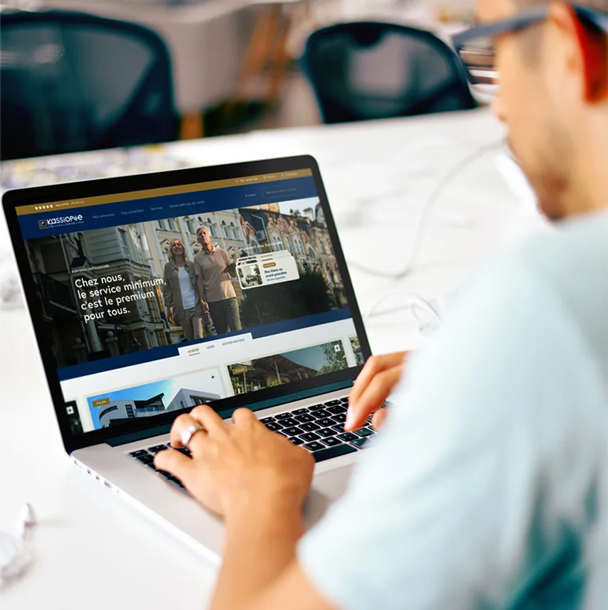 Personne utilisant un ordinateur portable affichant un site immobilier avec le texte « Chez nous, le service minimum, c'est le premium pour tous ».