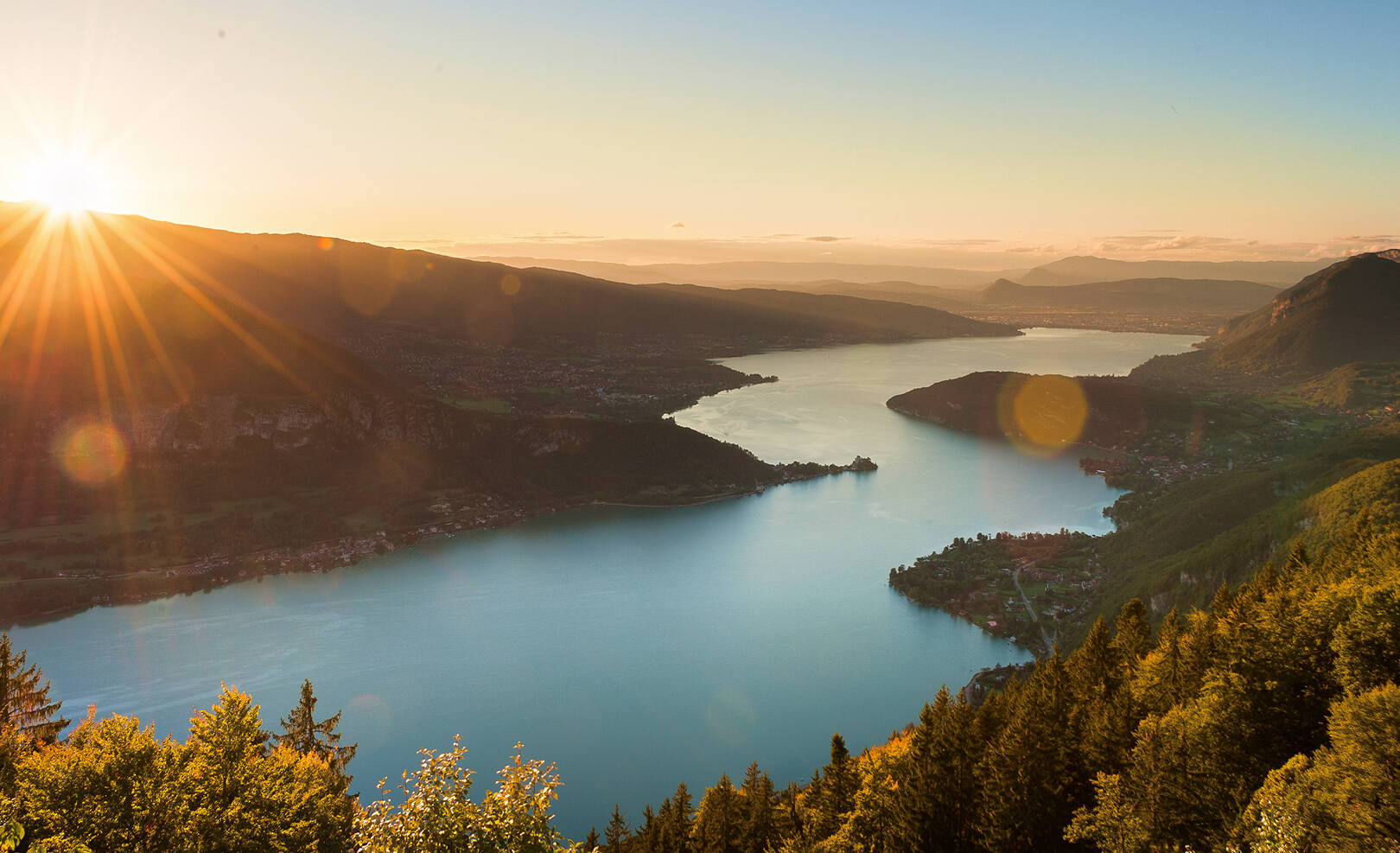 Coucher de soleil sur un lac entouré de montagnes et de forêts vertes.