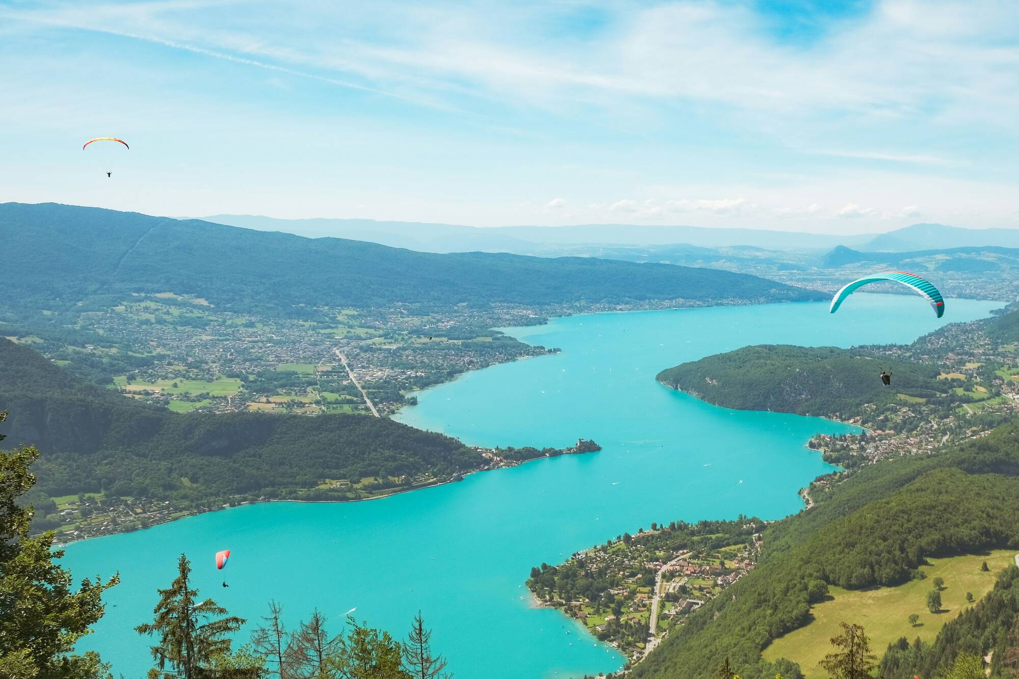Parapentes volant au-dessus d'un lac bleu entouré de montagnes et de villages verts.