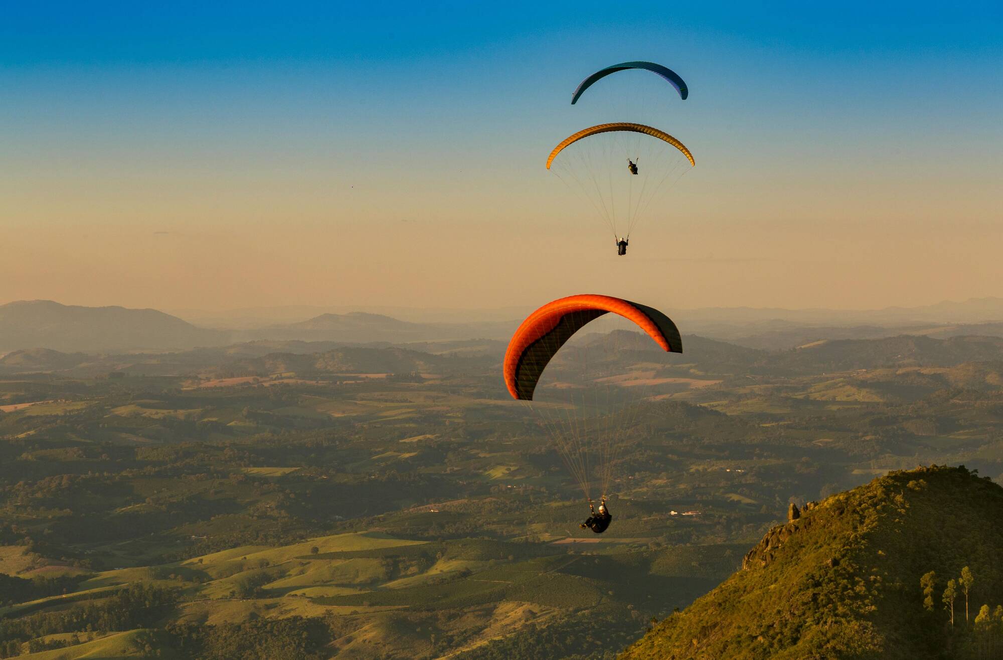 Trois parapentistes volant au-dessus de collines verdoyantes sous un ciel clair au coucher du soleil.