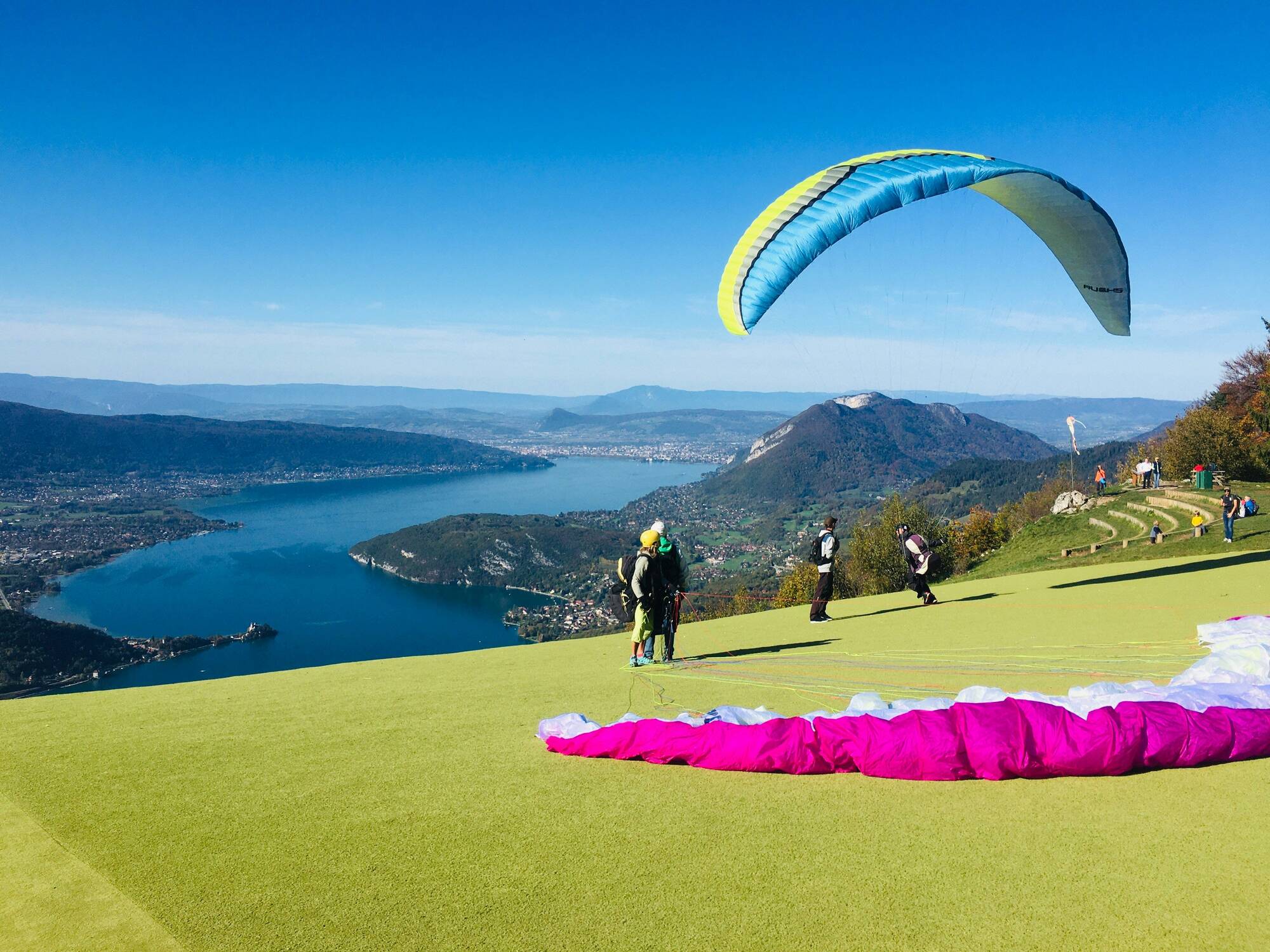 Parapentistes sur une colline avec vue sur un grand lac, montagnes et ciel bleu clair.