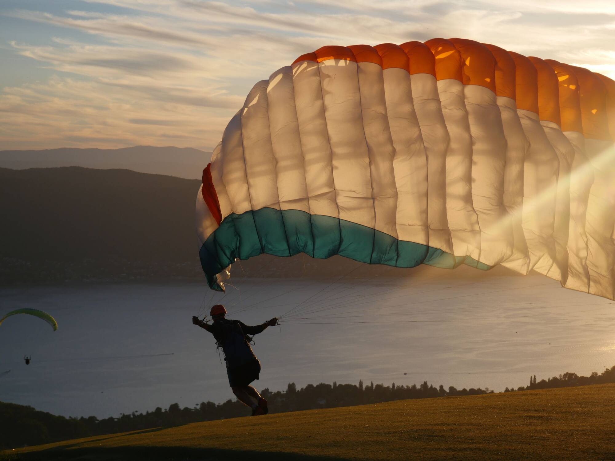 Parapentiste décolle sur une colline au coucher du soleil avec un parachute orange, blanc et bleu.