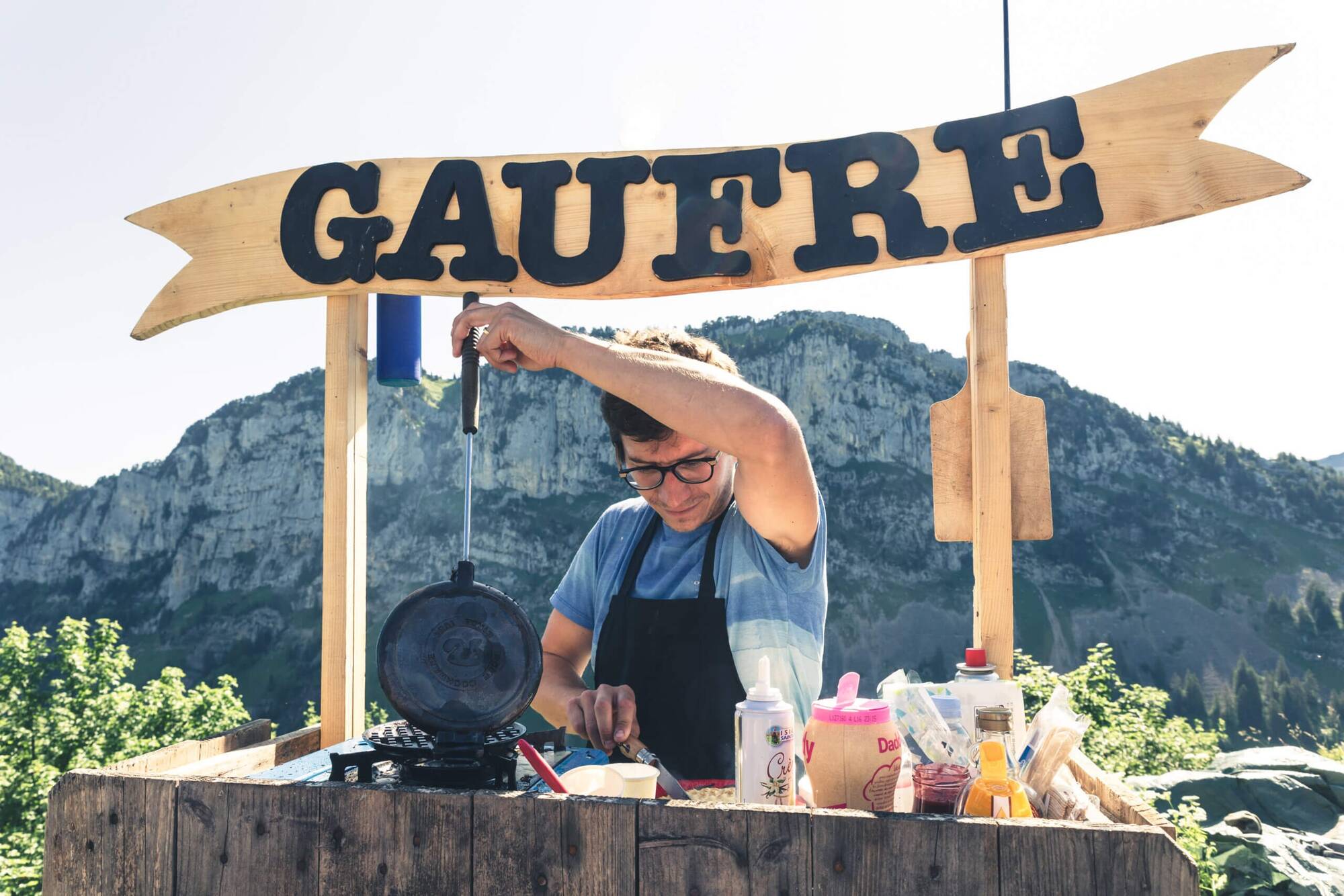 Un homme prépare des gaufres en plein air avec montagnes en arrière-plan.