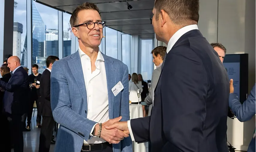 Two business leaders shaking hands at a Boardwave networking event with other attendees in a modern venue