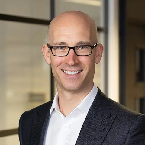 Smiling bald man wearing black glasses, white shirt, and dark suit jacket in an office setting.