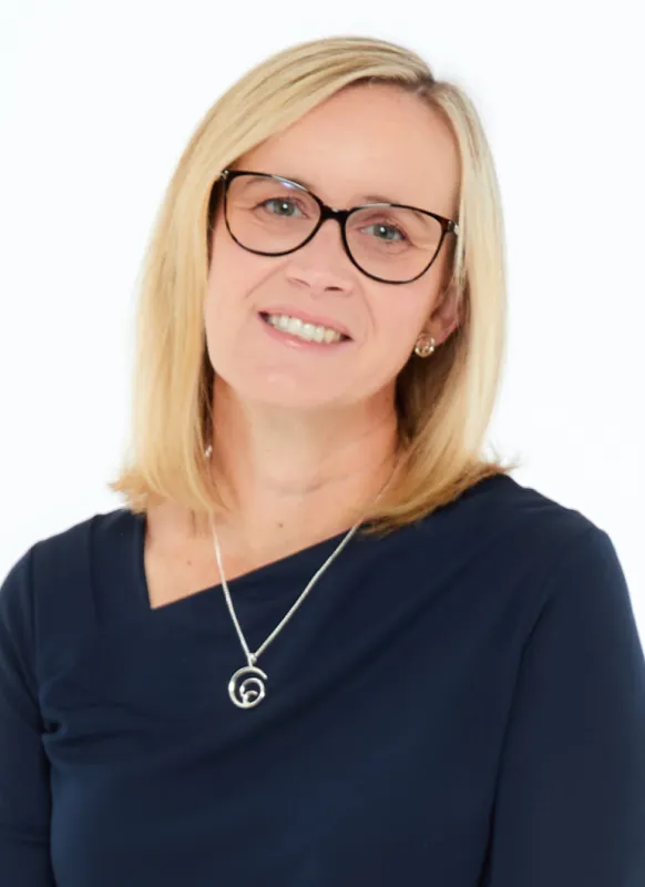 Smiling woman (Stephanie Hutton) with blonde hair wearing glasses, a navy blue top, and a silver necklace against a white background.