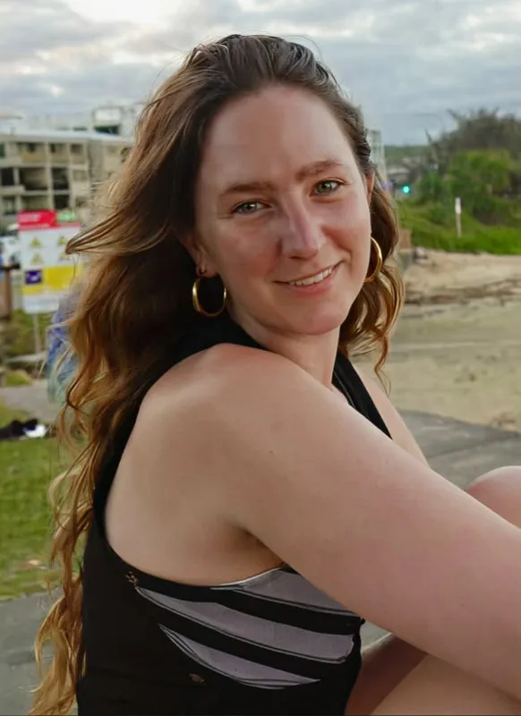 Smiling woman (Cat Trelawny) with long brown hair wearing hoop earrings and a black sleeveless top, sitting outdoors near a beach.