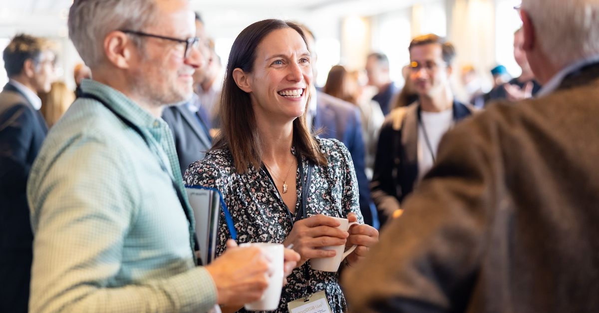 Three adults in conversation at a social or professional event, holding white coffee mugs, with other people blurred in the background.