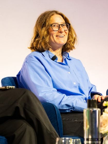 Person with shoulder-length wavy hair and glasses wearing a blue button-up shirt, sitting and smiling in a chair against a light background.