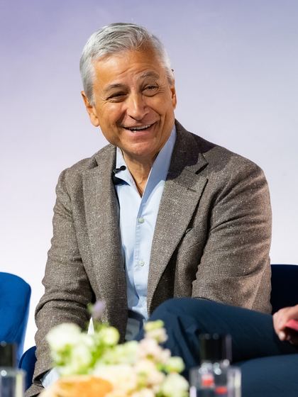 Smiling middle-aged man with gray hair wearing a brown blazer and blue shirt, sitting on a blue chair with a blurred flower arrangement in the foreground.