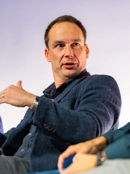 Man in a dark blazer speaking and gesturing with his hand during a discussion or panel event.