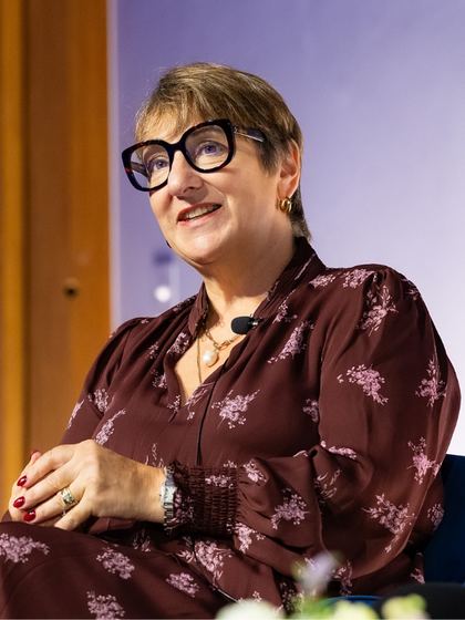 Woman with short hair and large black-framed glasses speaking, wearing a maroon floral dress and gold hoop earrings, seated with her hands resting on her lap.