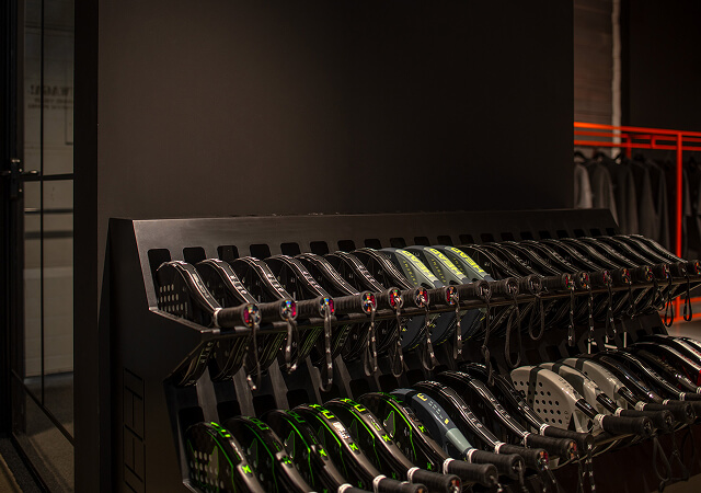 Rows of padel rackets neatly arranged on a black rack in a dimly lit sports facility.