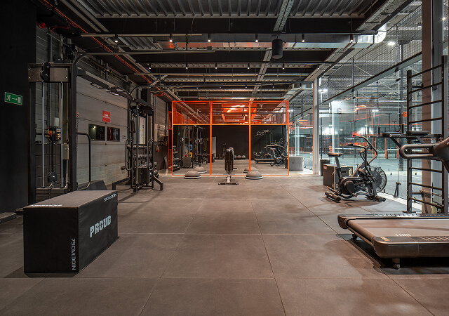 Modern gym space with exercise equipment including a treadmill, stationary bike, multi-gym station, and plyometric box, featuring an industrial design and orange-lit area in the background.