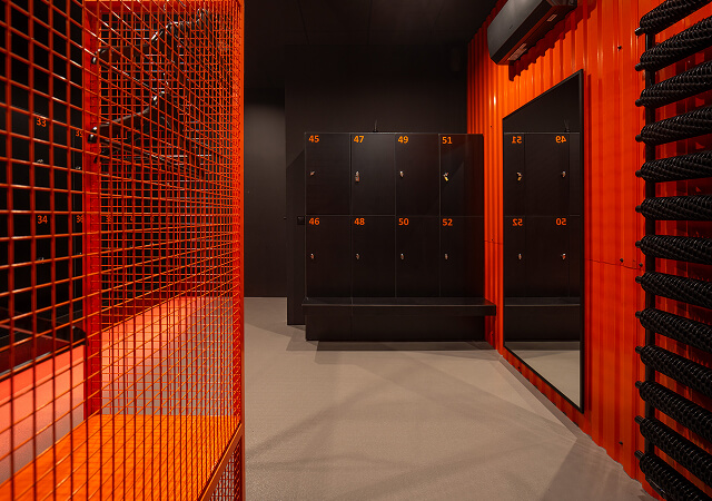 Locker room with numbered black lockers, an orange metal mesh bench, a large mirror, and a black wall-mounted air conditioner.