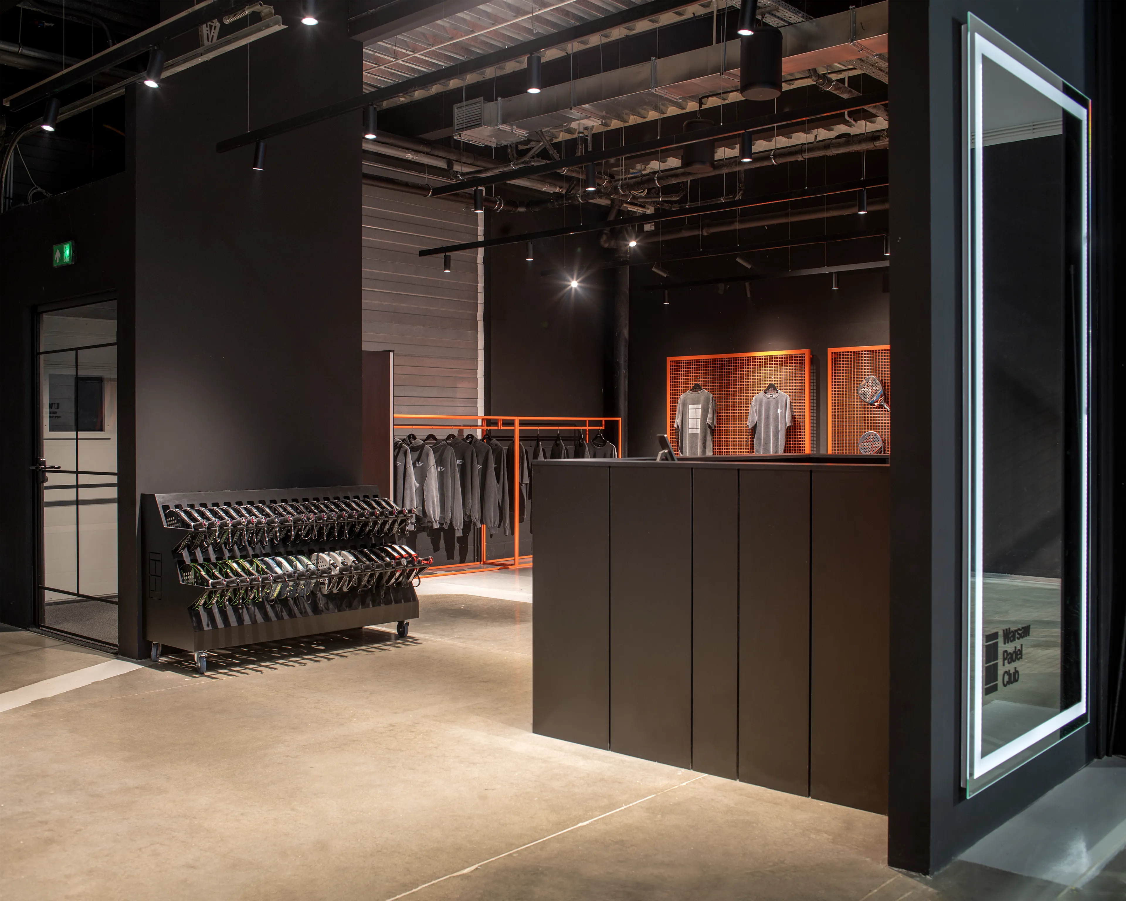 Modern facility reception area with black walls, a black counter, racks of hanging gray sweatshirts, and shelves displaying padel rackets.