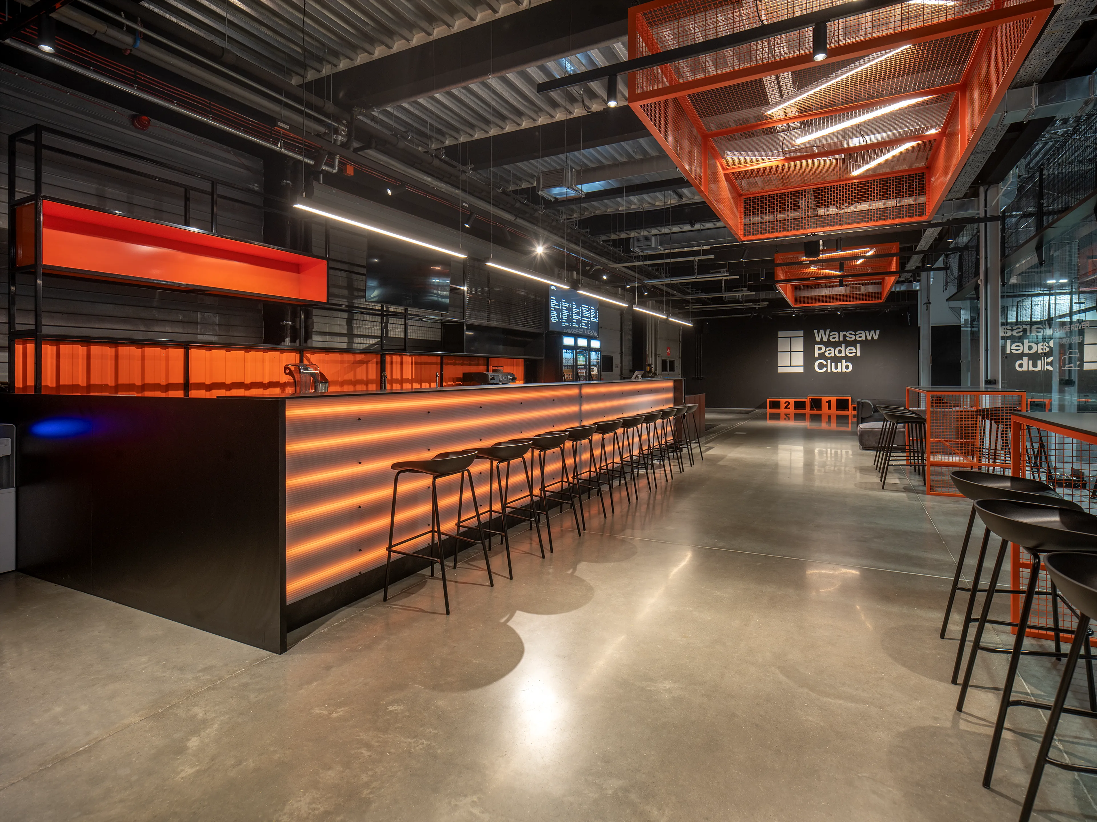 Modern bar area with orange illuminated front desk, black stools, and industrial ceiling at Warsaw Padel Club.