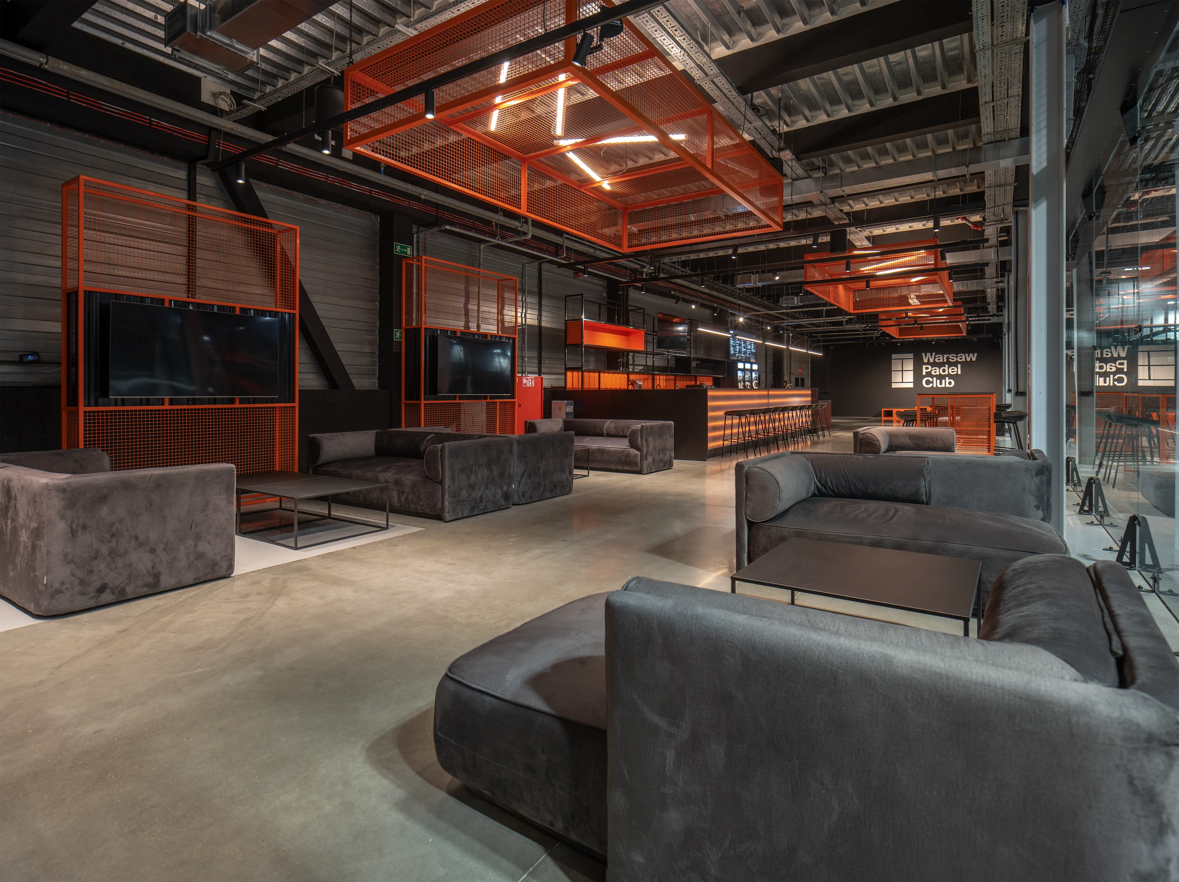 Modern lounge area with grey sofas, orange metal shelves, multiple flat screens, and a long bar under industrial ceiling fixtures.