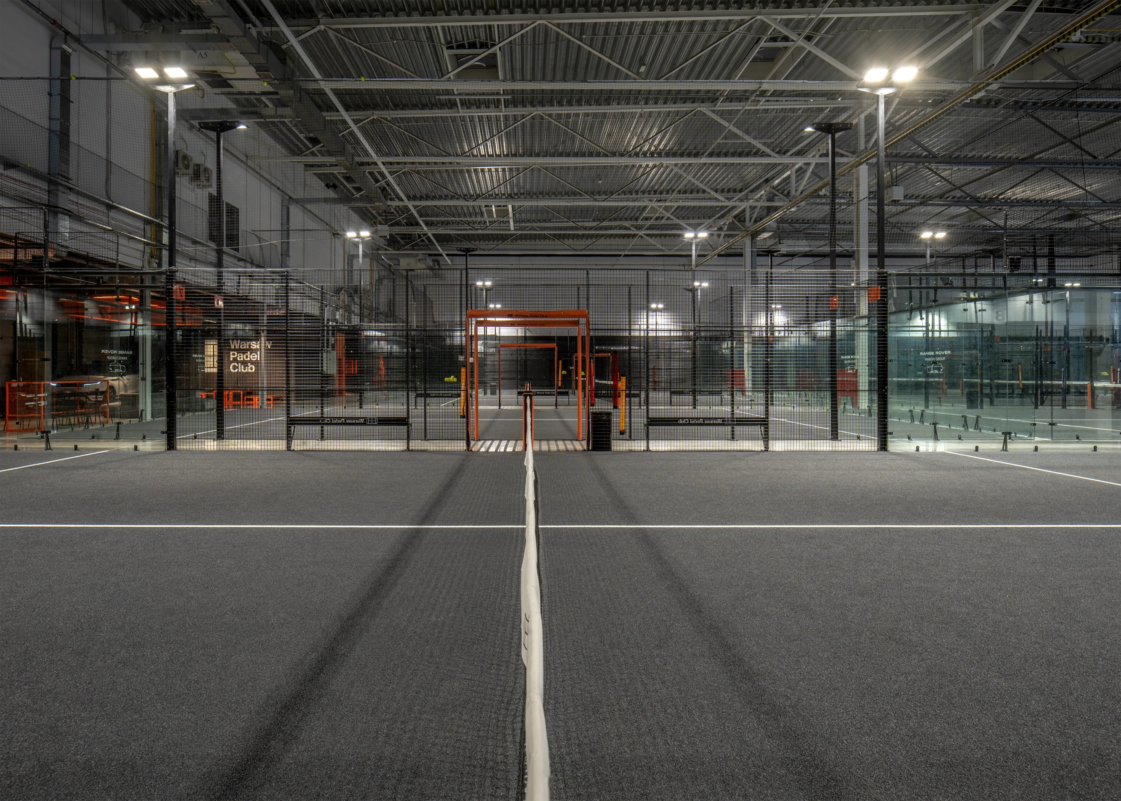 Indoor padel court with gray floor, white boundary lines, fenced walls, and bright overhead lights in a large sports facility.