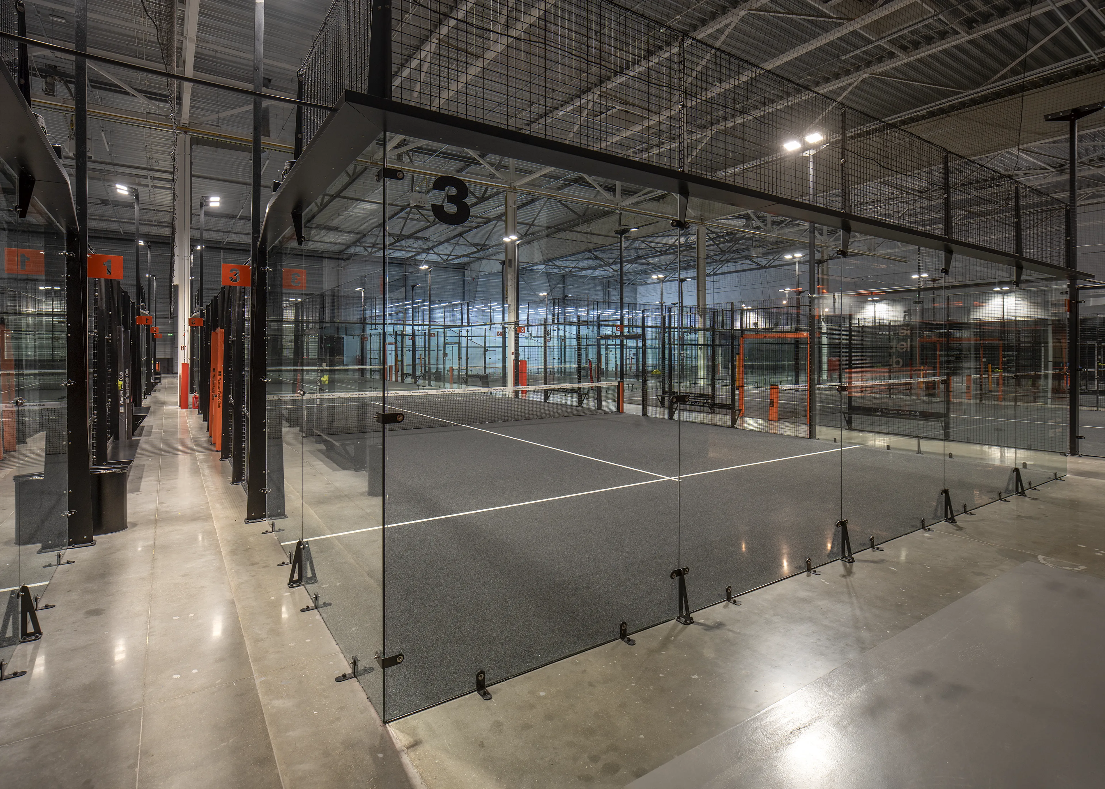 Indoor padel club with multiple enclosed glass courts numbered 1 to 3, featuring gray flooring and industrial lighting.