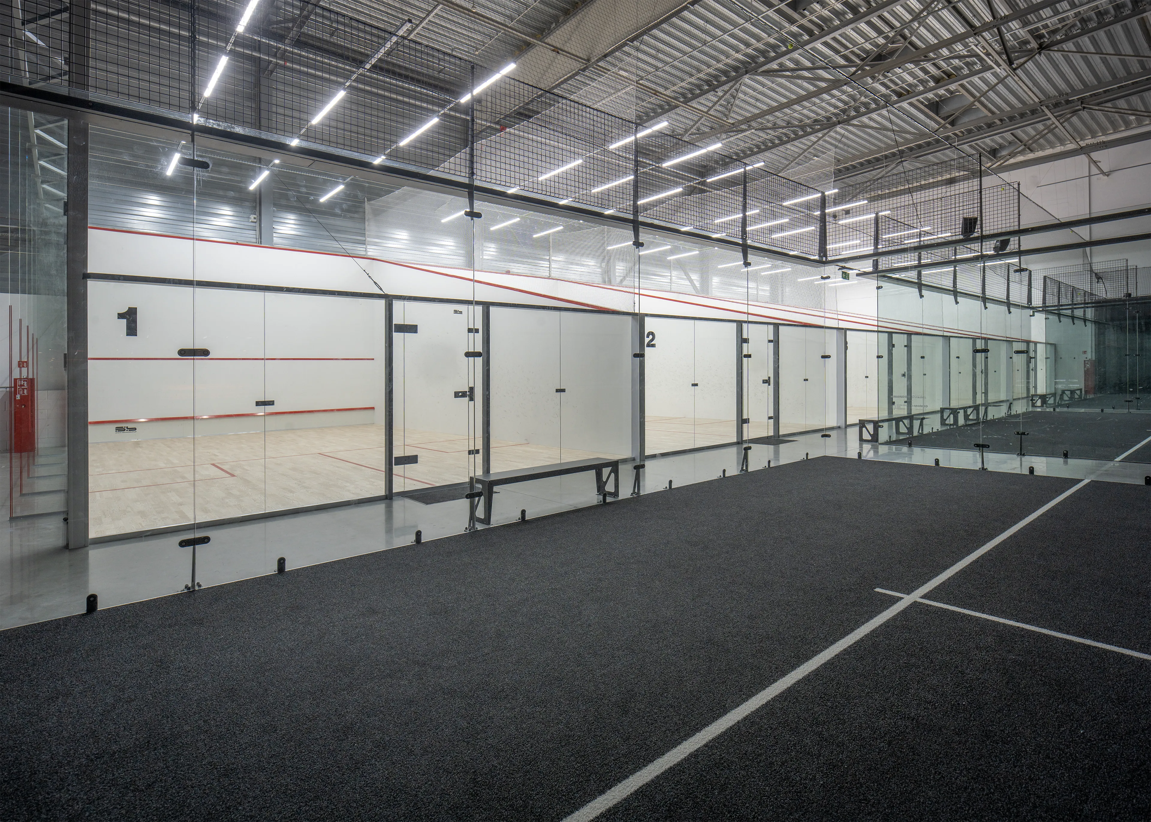 Indoor sports facility with glass-enclosed squash courts numbered 1 and 2, and a dark carpeted floor in the foreground.