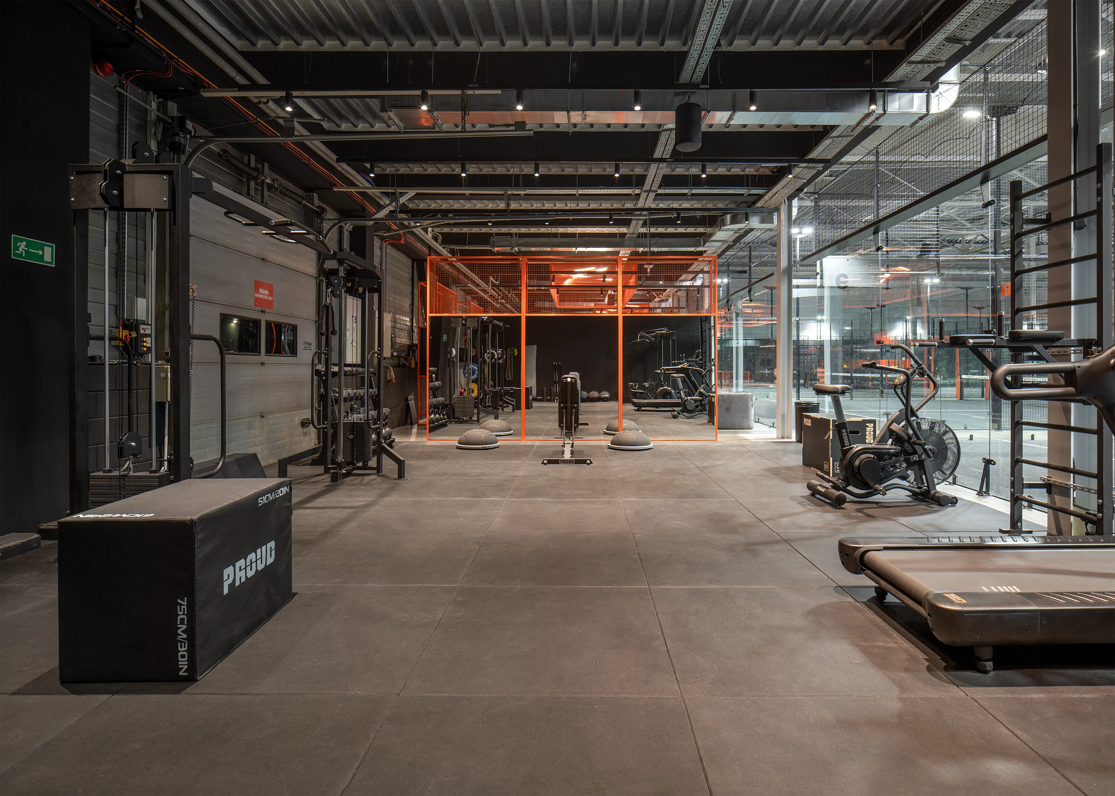 Modern gym interior with various fitness equipment including a padded box, treadmill, rowing machine, exercise bikes, and weight machines under an industrial ceiling.