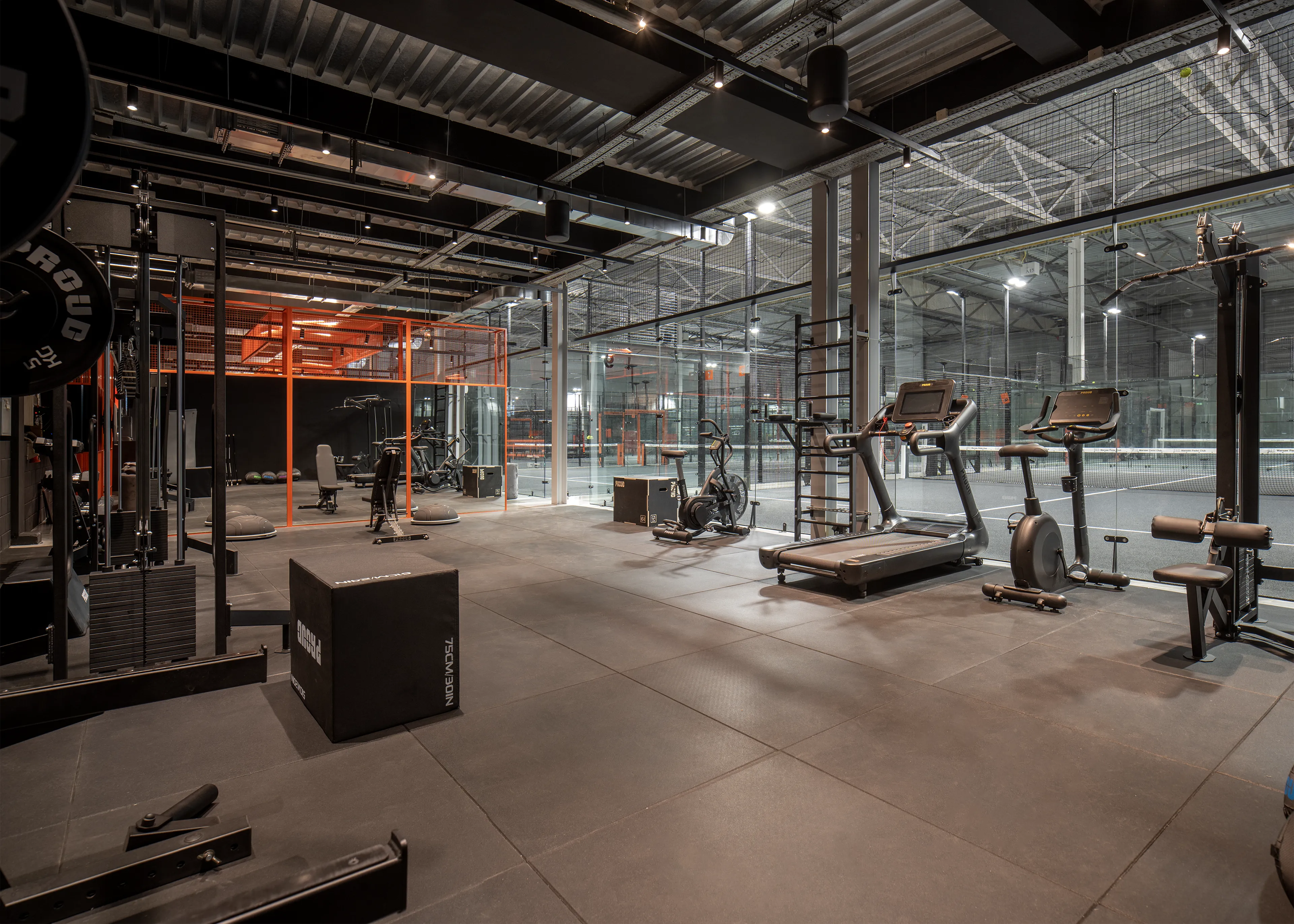 Modern gym with various exercise equipment including treadmills, stationary bikes, weight machines, and plyometric boxes, adjacent to glass padel courts.