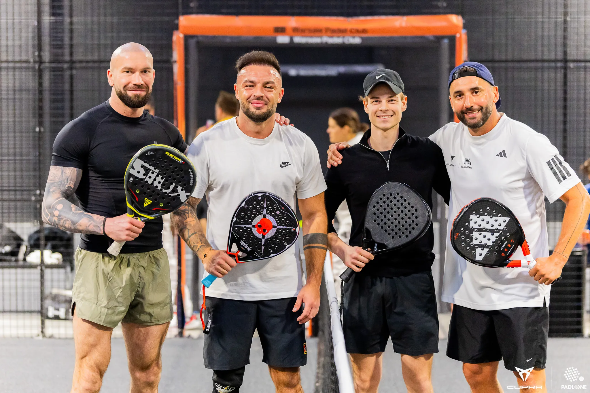 Four men standing on a padel court holding padel rackets, smiling at the camera.
