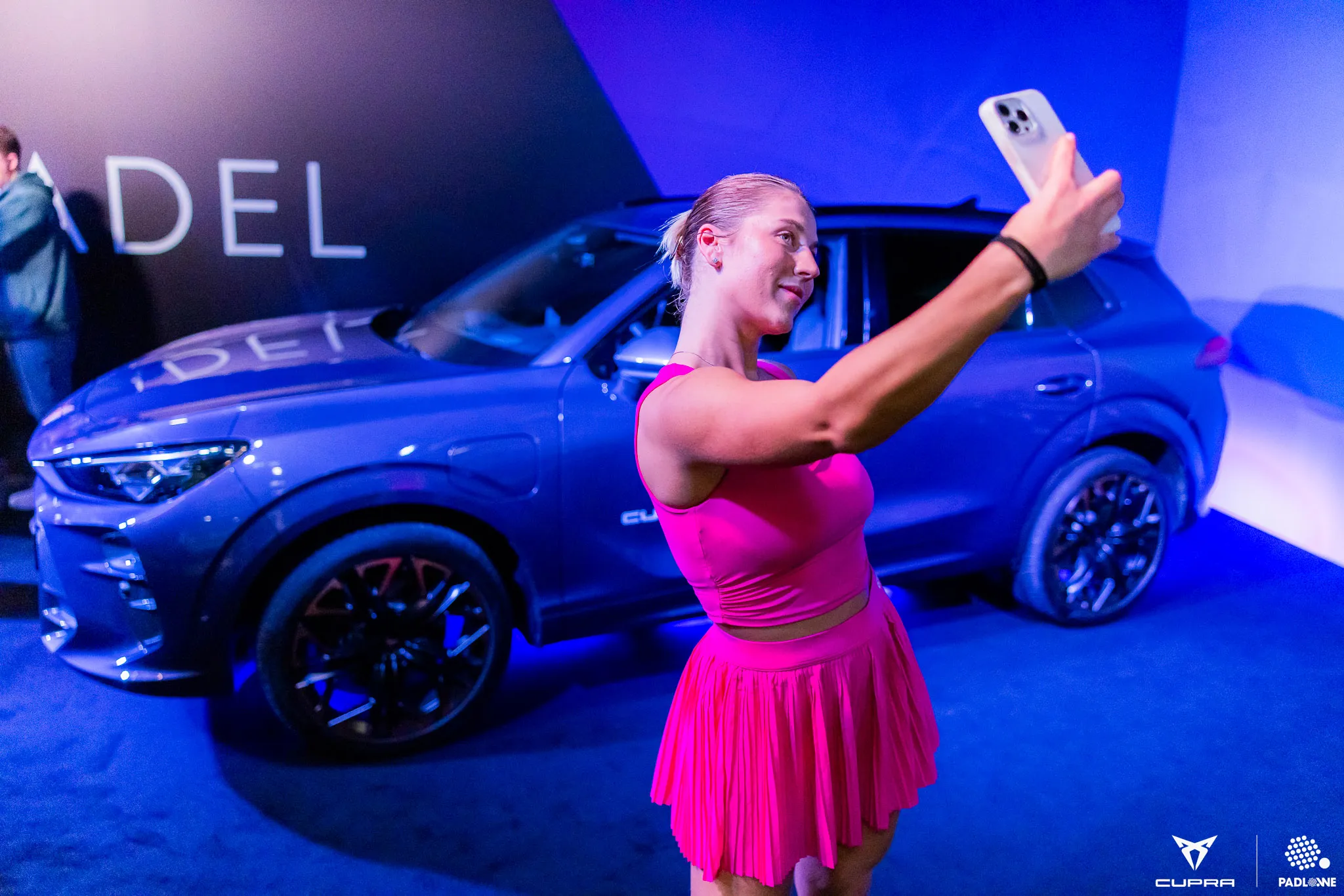 Woman in bright pink outfit taking a selfie with a gray CUPRA SUV in the background under blue lighting.