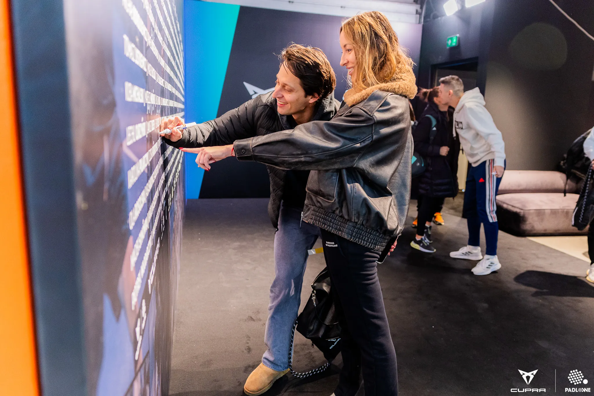 Two people smiling and writing on a large wall display indoors, with others in the background near a couch.