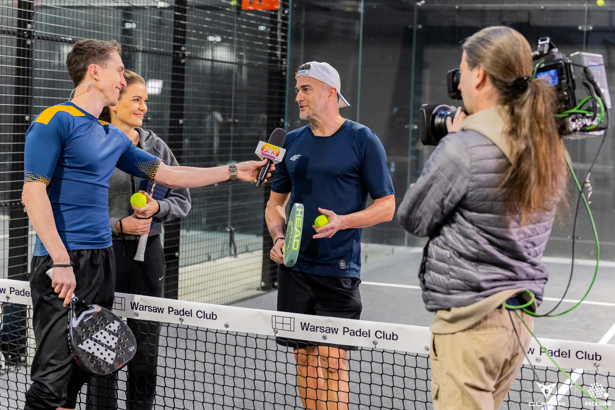 Two padel players being interviewed on court by a reporter holding a microphone, with a cameraman filming.