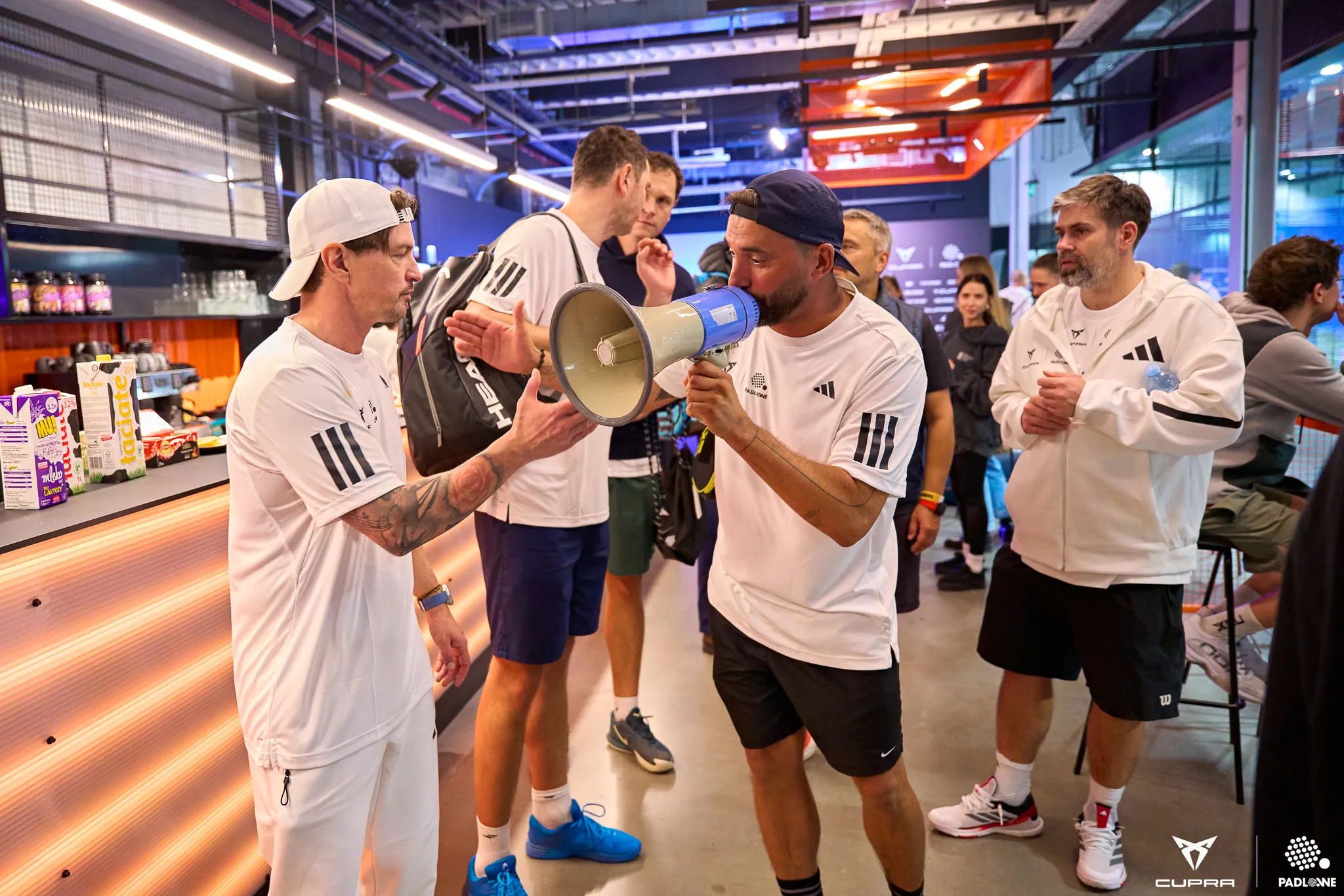 Group of men in white sportswear inside a modern indoor space, one speaking into a megaphone while others listen and interact.