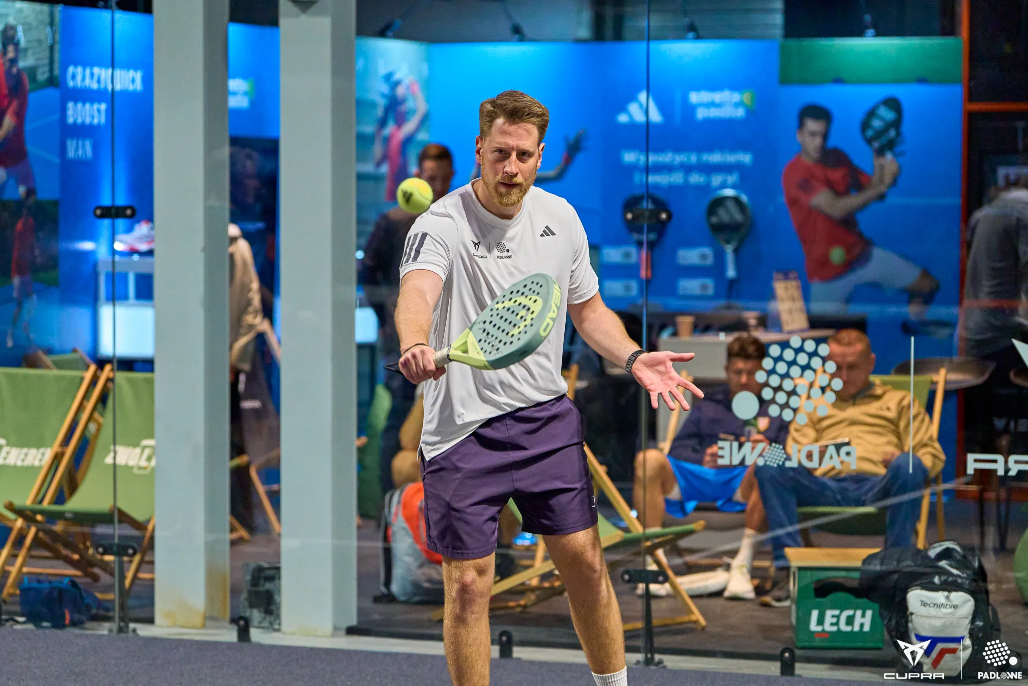 Man in white shirt and purple shorts playing padel inside a court, holding a padel racket and focusing on a yellow ball in midair.