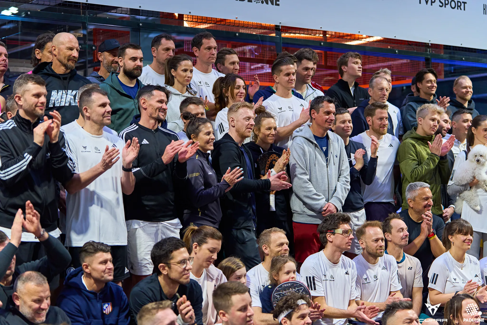 Large group of men and women in sportswear clapping and posing indoors, some holding padel rackets and a small white dog.