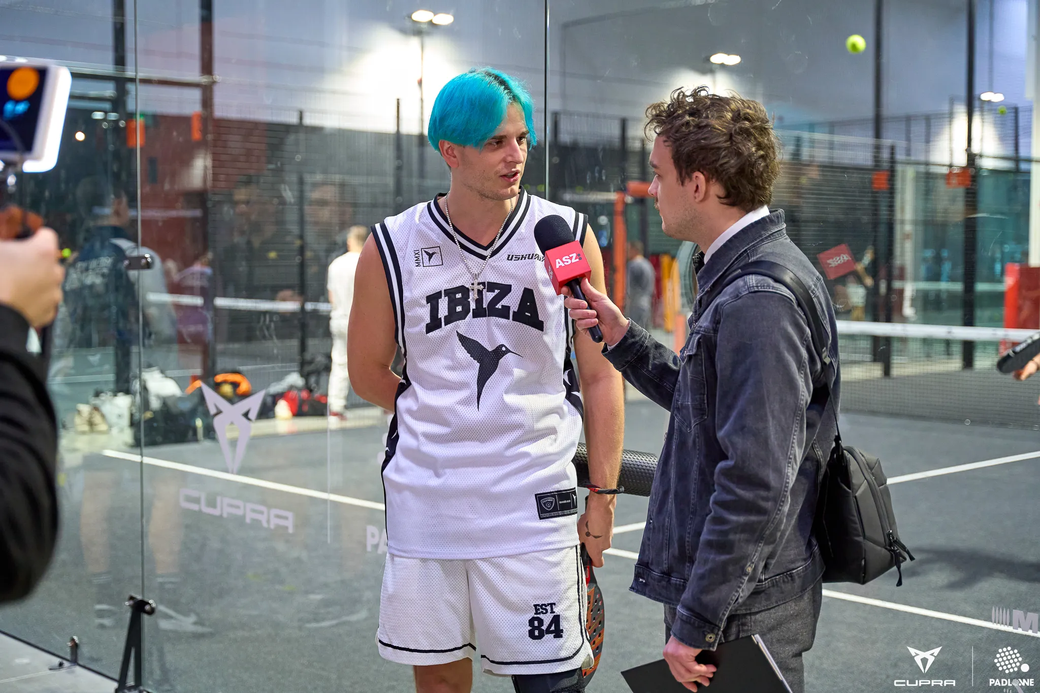 Man with blue hair wearing an Ibiza sports jersey being interviewed on a padel court.