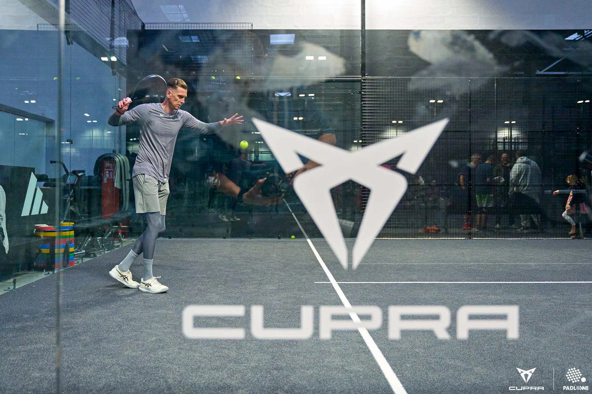 Man in athletic wear playing padel on an indoor court with large CUPRA logo on the glass wall.