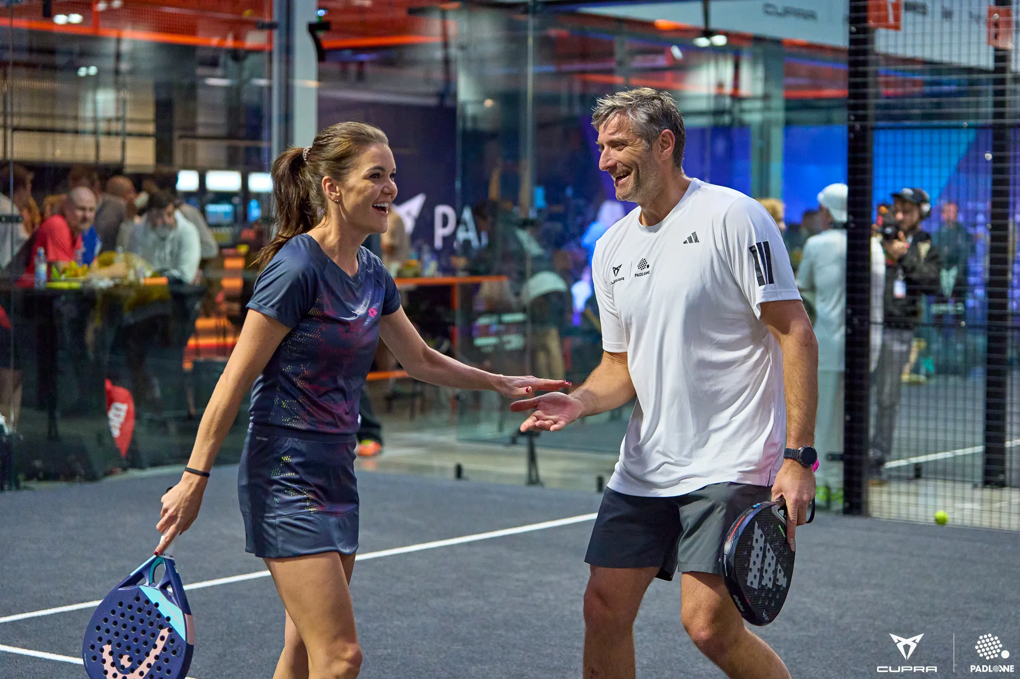 A man and woman smiling and shaking hands on an indoor padel court, both holding padel rackets.