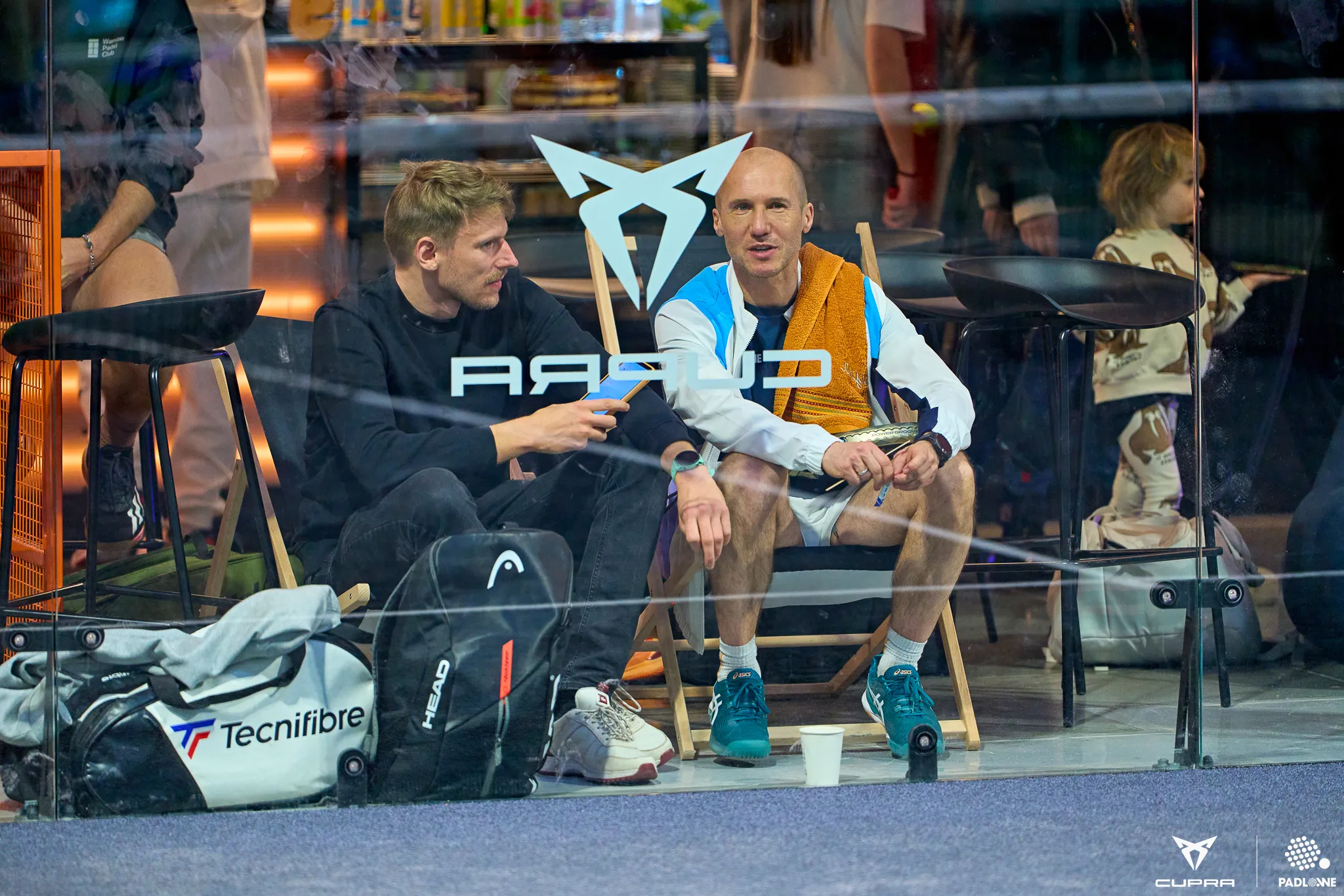 Two men sitting on chairs behind glass with sports bags on the floor, one man holds a padel racket, a child stands in the background.