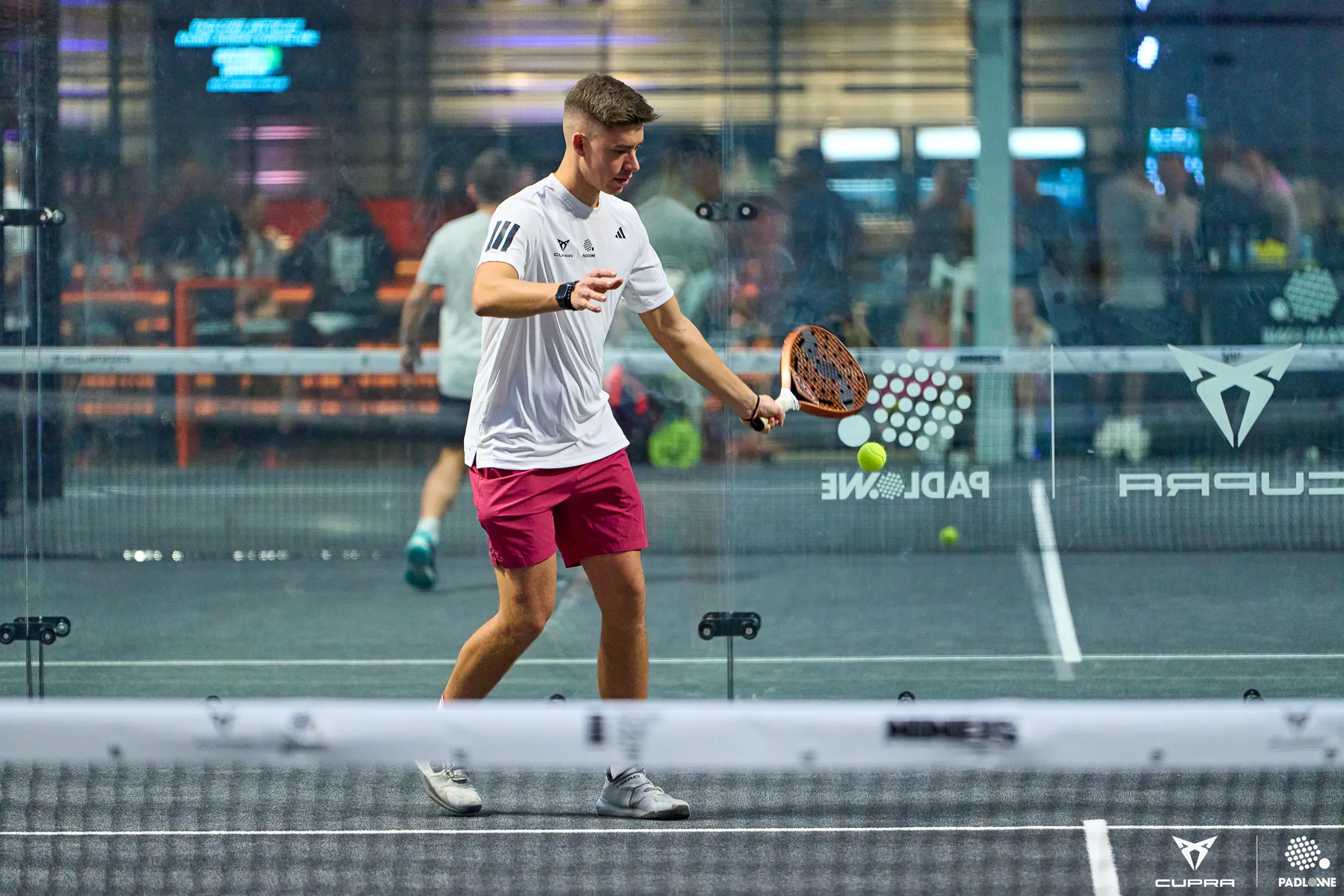 Young man in pink shorts and white shirt playing padel, hitting a ball with a paddle inside a glass court.