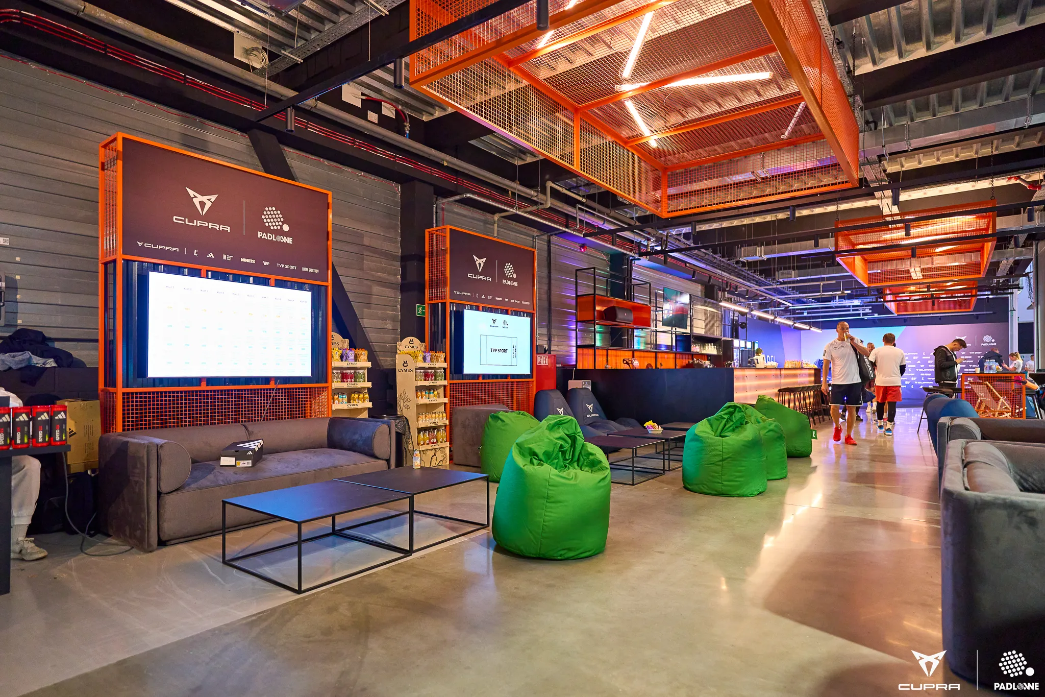 Modern indoor lounge area with gray sofas, green bean bags, coffee tables, large digital screens with CUPRA and PADLOWE logos, and people walking near a bar.