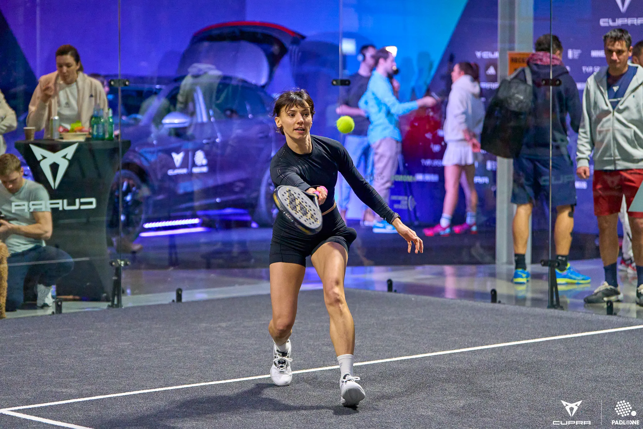 Female padel player in black sportswear lunging forward to hit a ball on an indoor court with spectators and promotional banners in the background.