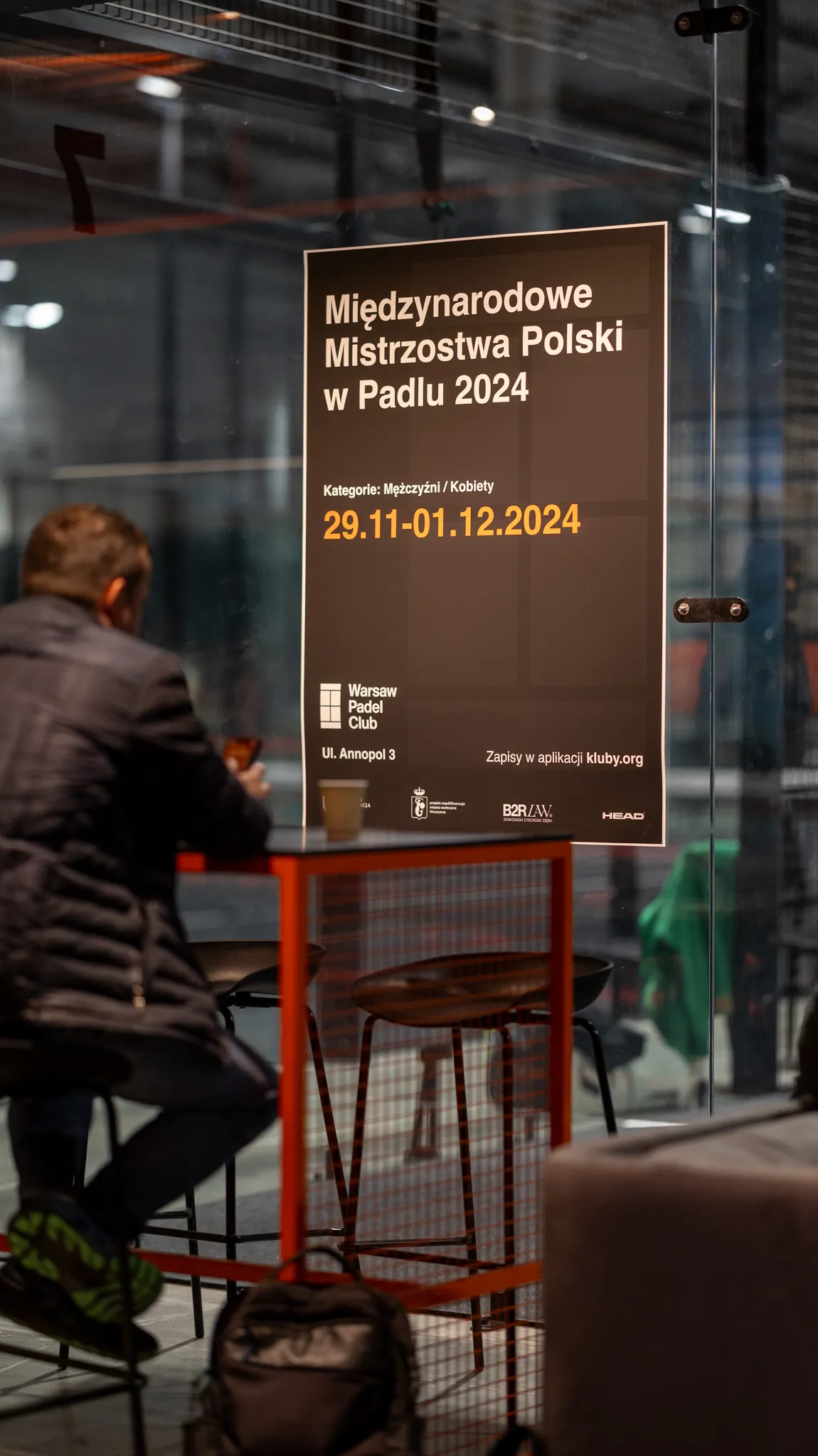 Man in a jacket sitting at a red table inside a glass-walled padel club near a poster advertising the 2024 International Polish Padel Championships from November 29 to December 1.