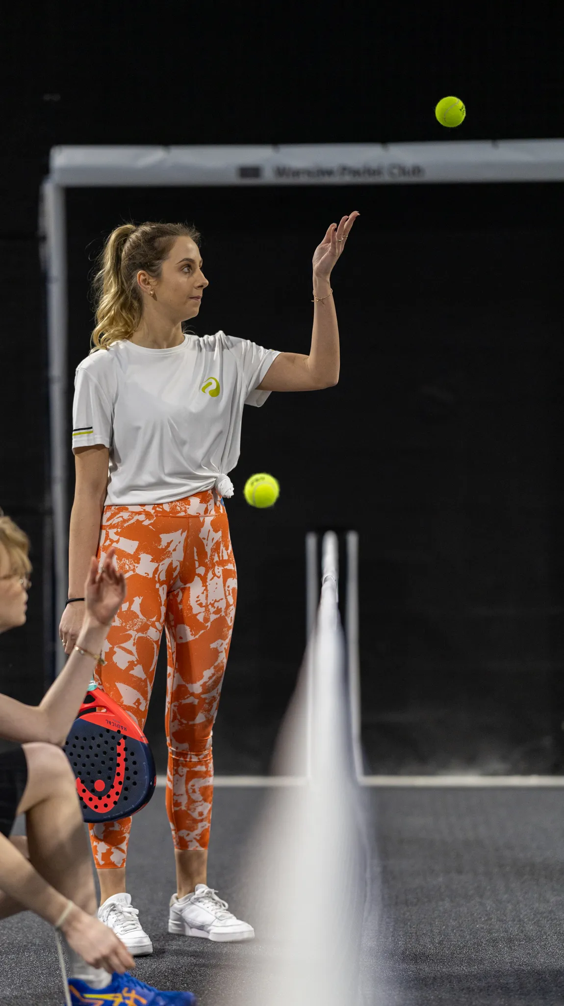 Woman in white shirt and orange patterned leggings tossing a yellow padel ball on a padel court.