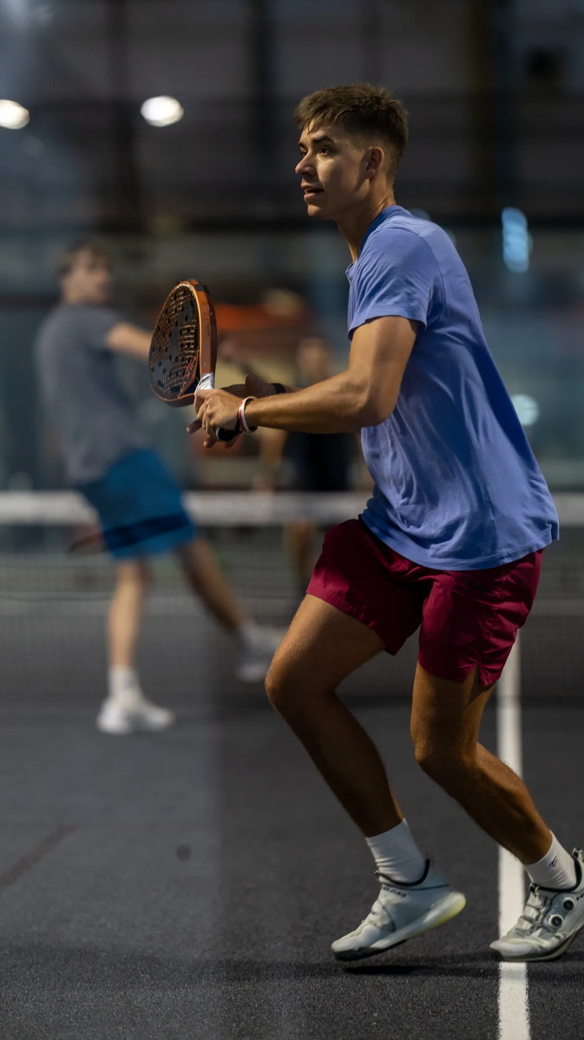 Young man in blue shirt and maroon shorts playing padel indoors, holding a padel racket and preparing to hit the ball.