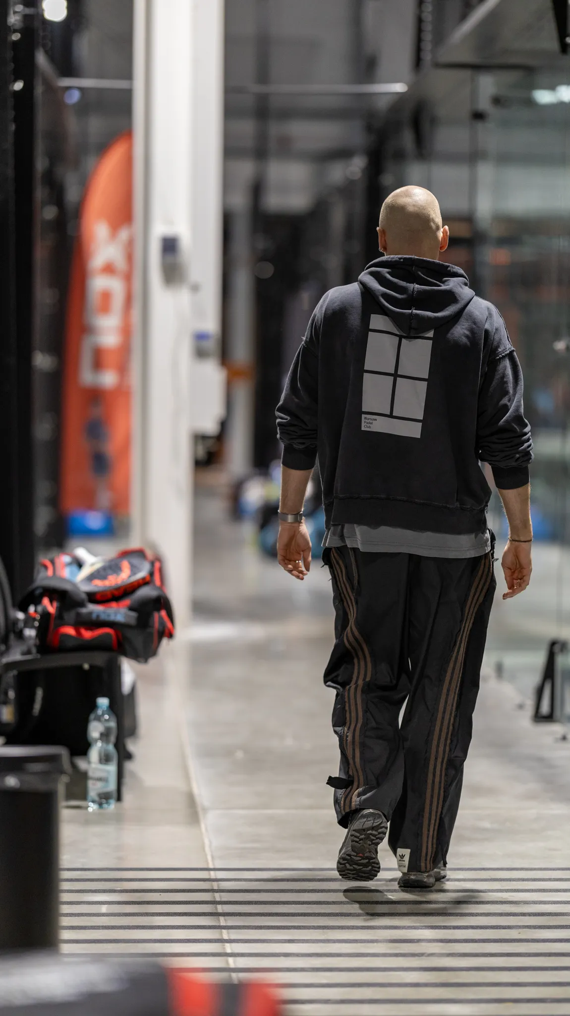 Man with shaved head walking away indoors wearing black hoodie with grid design and black track pants with brown stripes.